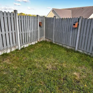 Backyard corner enclosed by a grey wooden fence, with grass and birdhouses, in preparation for building a DIY natural pond