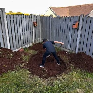 Farah digging the foundation for a DIY natural pond in a backyard corner enclosed by a grey wooden fence