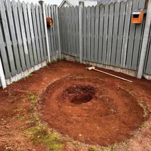 Completed excavation for a DIY natural pond in a backyard, with a tool resting beside the dug-out area