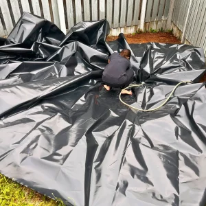 Farah positioning a black pond liner over the excavated area in a backyard for a DIY natural pond